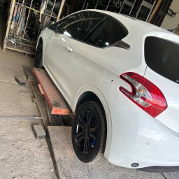 MOTOINVEST Paweł Nowrotek - Biały samochód na podnośniku w warsztacie, widoczne czarne felgi i tylne światło, w tle opony i wyposażenie warsztatowe.