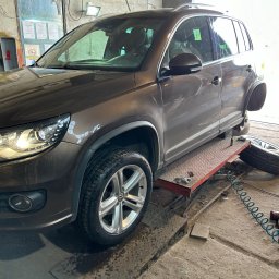 MOTOINVEST Paweł Nowrotek - Brązowy samochód osobowy na podnośniku w warsztacie, jedno koło zdjęte, obok leży opona, widoczny pistolet pneumatyczny z niebieskim wężem. Zakaz wstępu na ścianie.