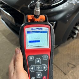 MOTOINVEST Paweł Nowrotek - Diagnostyka czujnika ciśnienia w oponach za pomocą urządzenia Autel MaxiTPMS TS508, widoczny fragment samochodu w tle.