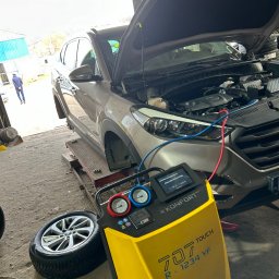 MOTOINVEST Paweł Nowrotek - Szary SUV na podnośniku w warsztacie z otwartą maską, podłączony do żółtego urządzenia diagnostycznego TEXA KONFORT 707 TOUCH R-1234 YF, widoczne manometry i ekran urządzenia, obok leży opona...
