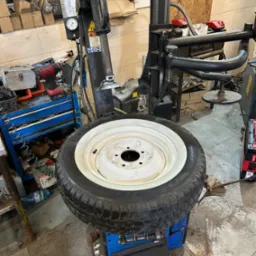 Opona z białym rantem osadzona na montażownicy do opon w warsztacie samochodowym, widoczne narzędzia i akcesoria warsztatowe w tle.