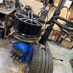 MOTOINVEST Paweł Nowrotek - Wymiana opony na maszynie Hofmann Monty 3300-22 SmartSpeed w warsztacie, felga aluminiowa z oponą na uchwycie, w tle narzędzia i skrzynki z częściami.