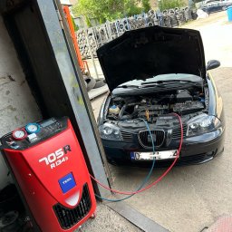 MOTOINVEST Paweł Nowrotek - Czarny samochód z otwartą maską podłączony do czerwonego urządzenia TEXA 705R R134a w warsztacie w Wałbrzychu, w tle sterta opon i budynek.