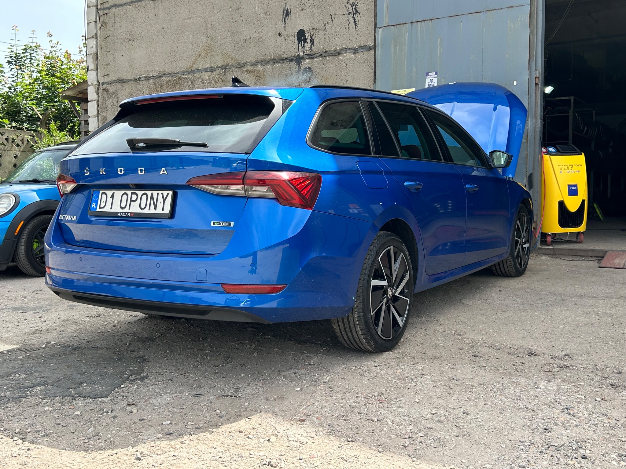 Niebieska Skoda Octavia kombi z otwartą maską stoi przed warsztatem; w tle widoczny żółty automat do obsługi klimatyzacji samochodowej Texa 707 Touch R-1234YF.