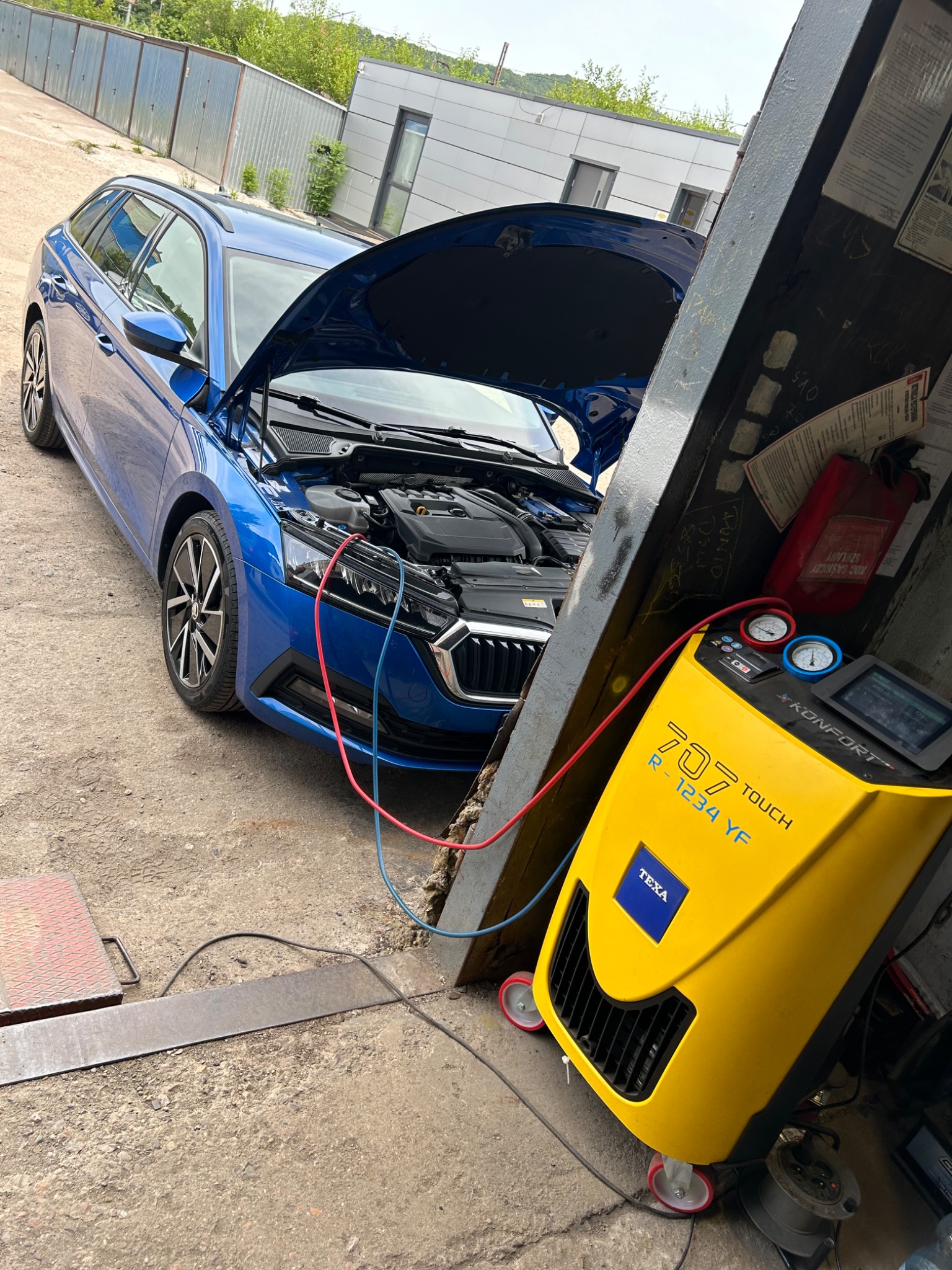 Niebieski samochód kombi z otwartą maską, podłączony przewodami do żółtego urządzenia TEXA Konfort 707 Touch do obsługi układów klimatyzacji w warsztacie samochodowym.