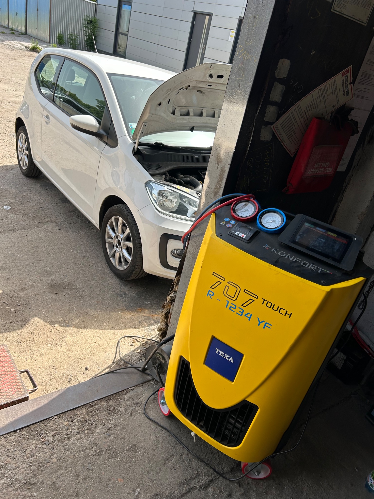 Biały samochód z otwartą maską w warsztacie, obok żółte urządzenie Texa 707 Touch R-1234 YF do serwisowania klimatyzacji, widoczne manometry i wyświetlacz.