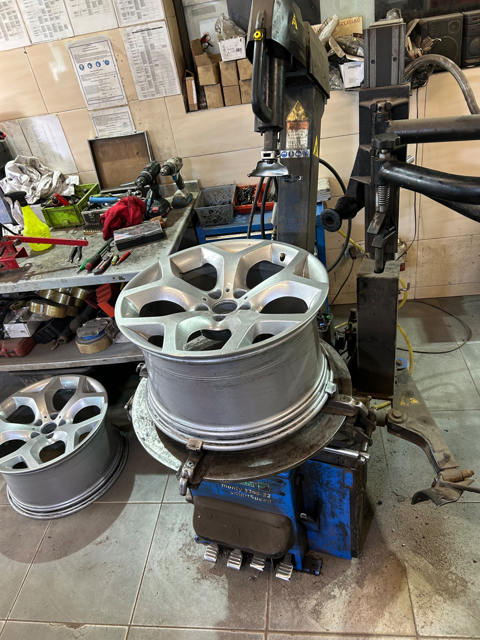 Aluminiowa felga na montażownicy w warsztacie, druga felga obok, narzędzia i papiery na ścianie w tle.