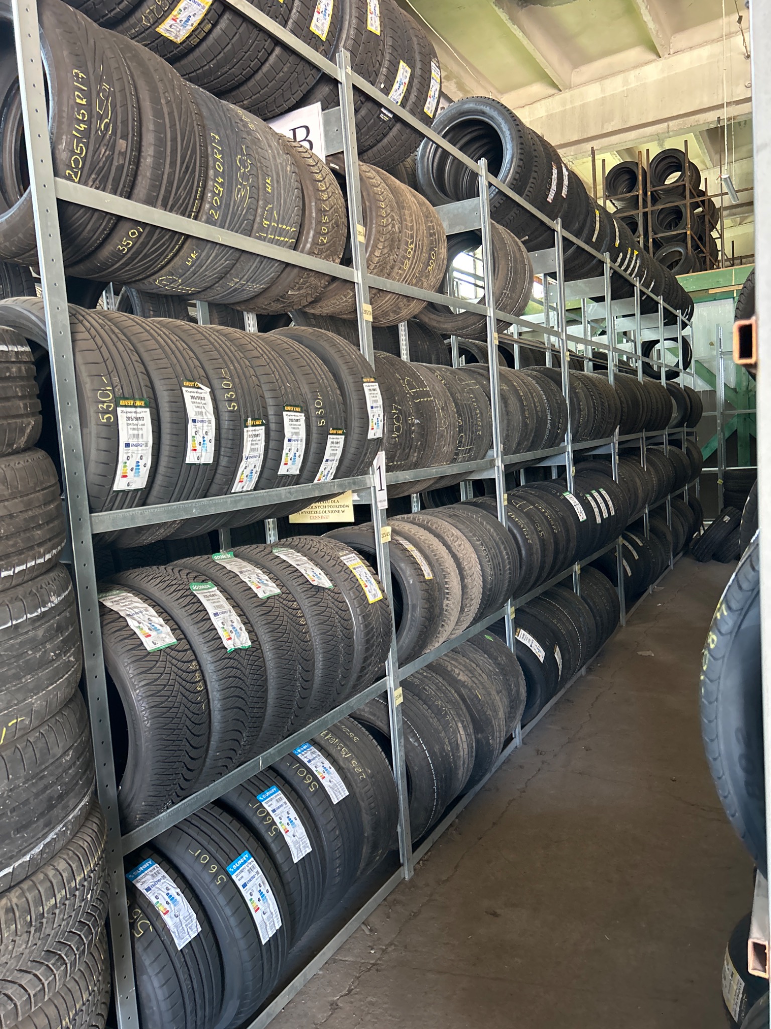 Magazyn opon samochodowych z różnymi modelami i rozmiarami ułożonymi na metalowych regałach, widoczne etykiety z parametrami opon.