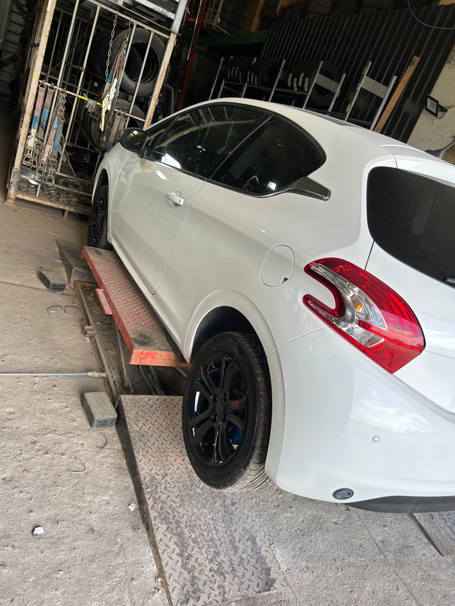 Biały samochód na podnośniku w warsztacie, widoczne czarne felgi i tylne światło, w tle opony i wyposażenie warsztatowe.