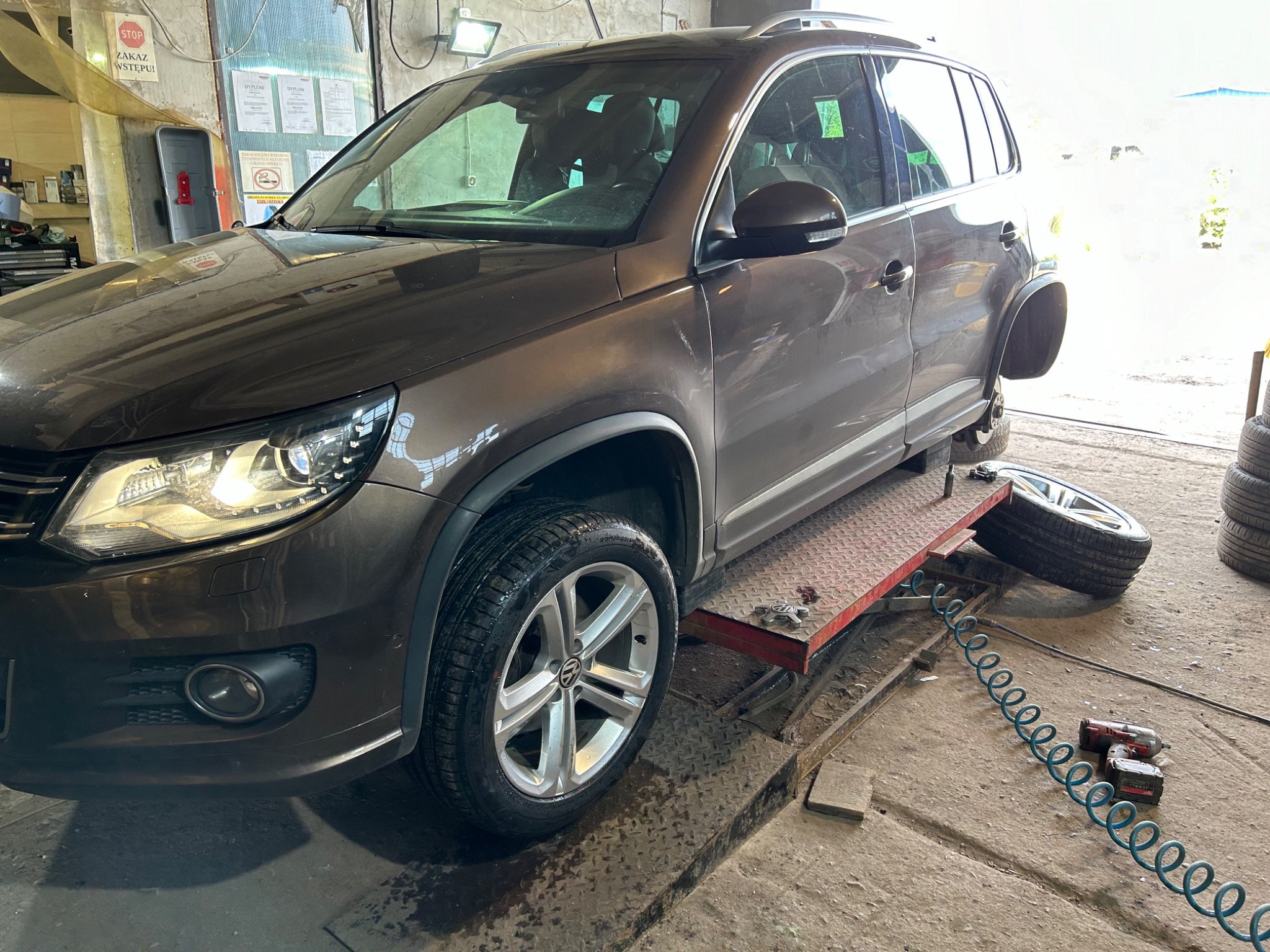 Brązowy samochód osobowy na podnośniku w warsztacie, jedno koło zdjęte, obok leży opona, widoczny pistolet pneumatyczny z niebieskim wężem. Zakaz wstępu na ścianie.