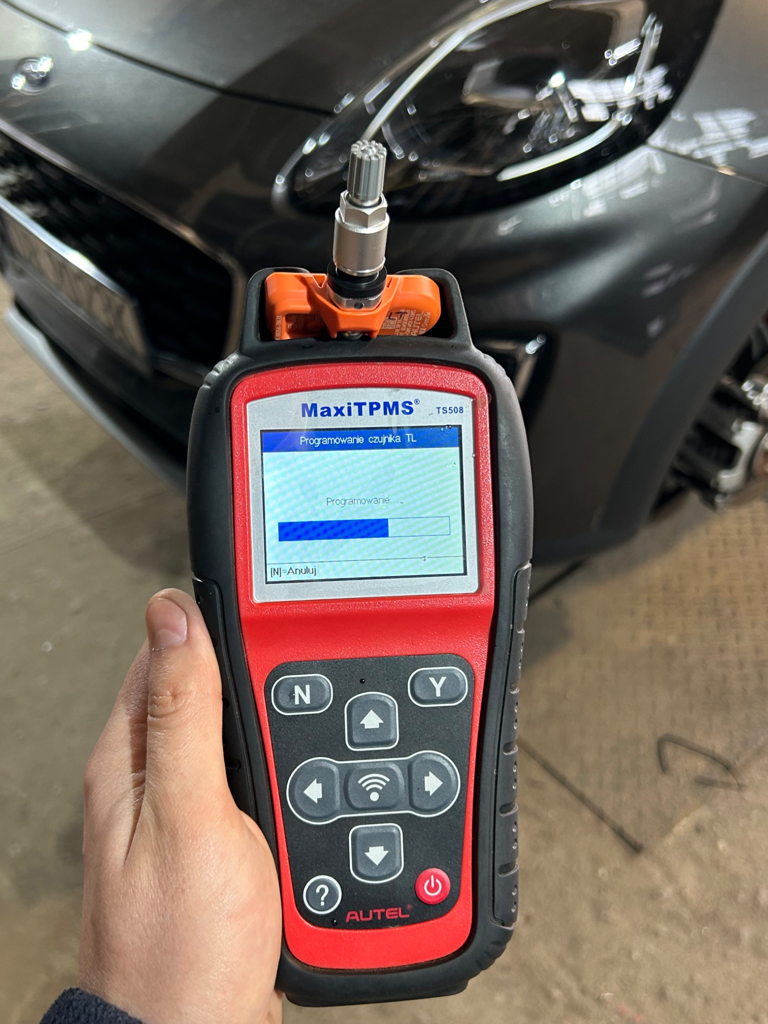 Diagnostyka czujnika ciśnienia w oponach za pomocą urządzenia Autel MaxiTPMS TS508, widoczny fragment samochodu w tle.
