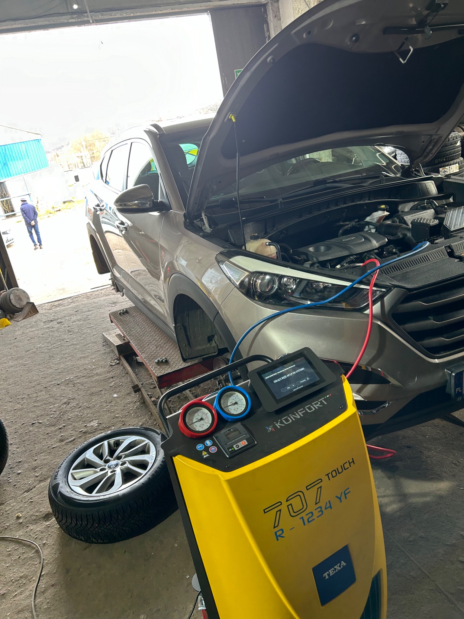 Szary SUV na podnośniku w warsztacie z otwartą maską, podłączony do żółtego urządzenia diagnostycznego TEXA KONFORT 707 TOUCH R-1234 YF, widoczne manometry i ekran urządzenia, obok leży opona...