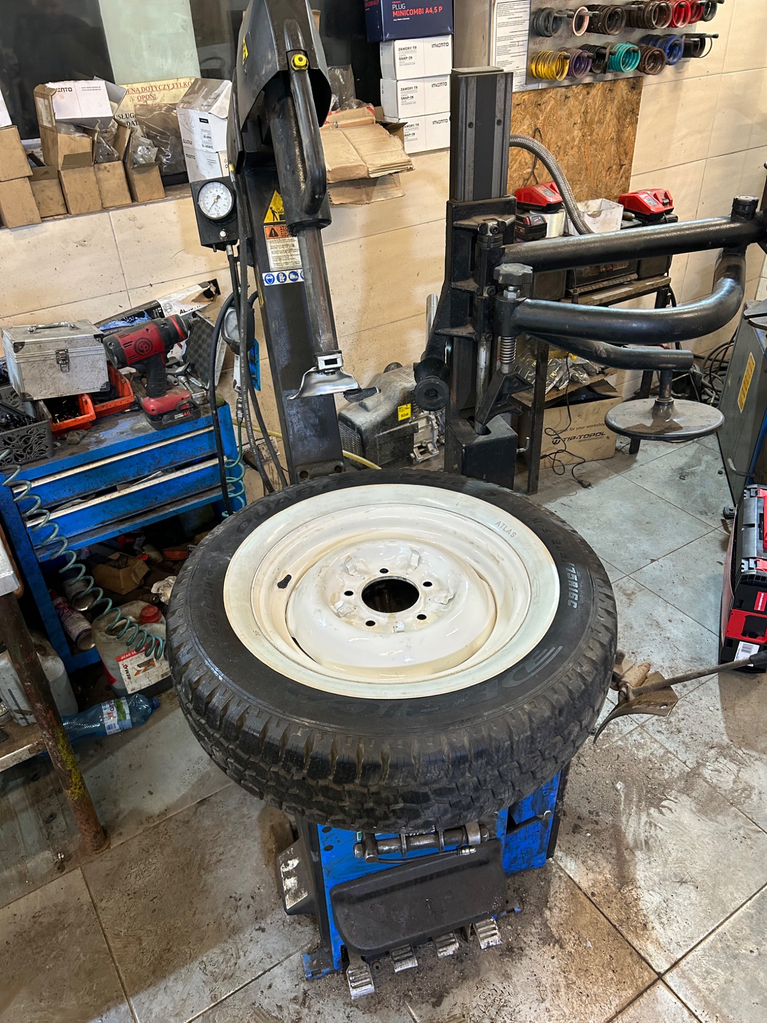 Opona z białym rantem osadzona na montażownicy do opon w warsztacie samochodowym, widoczne narzędzia i akcesoria warsztatowe w tle.