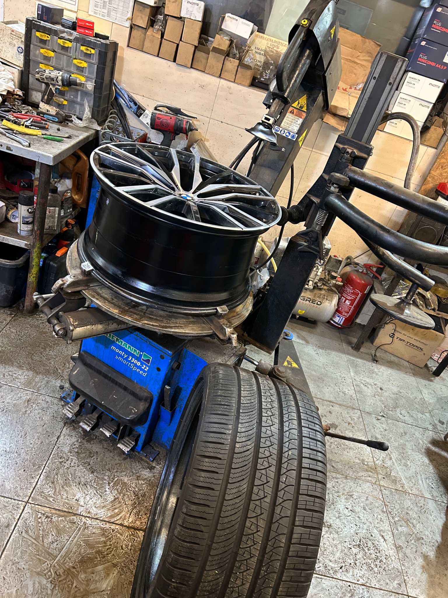 Wymiana opony na maszynie Hofmann Monty 3300-22 SmartSpeed w warsztacie, felga aluminiowa z oponą na uchwycie, w tle narzędzia i skrzynki z częściami.