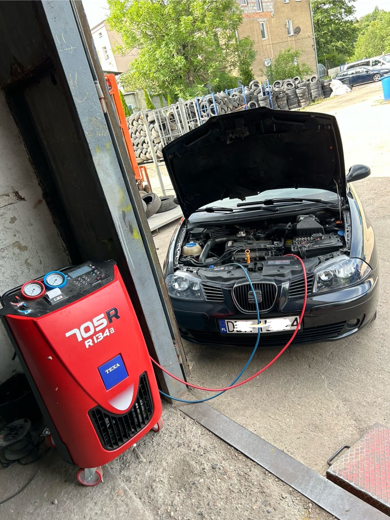 Czarny samochód z otwartą maską podłączony do czerwonego urządzenia TEXA 705R R134a w warsztacie w Wałbrzychu, w tle sterta opon i budynek.