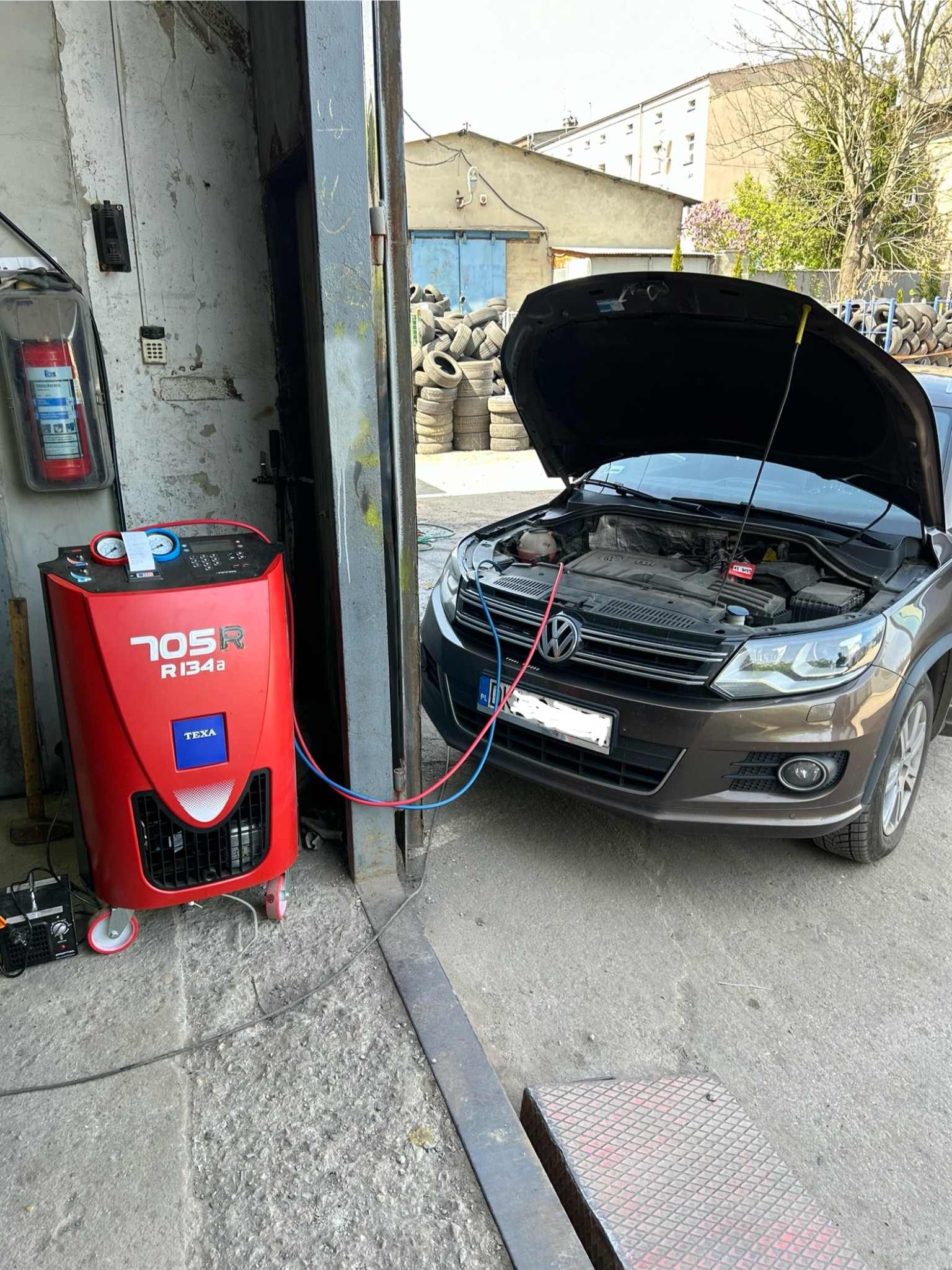 Brązowy samochód z otwartą maską podłączony do czerwonego urządzenia TEXA 705R R134a w warsztacie, z widocznymi przewodami, z tyłu stos opon i budynki.