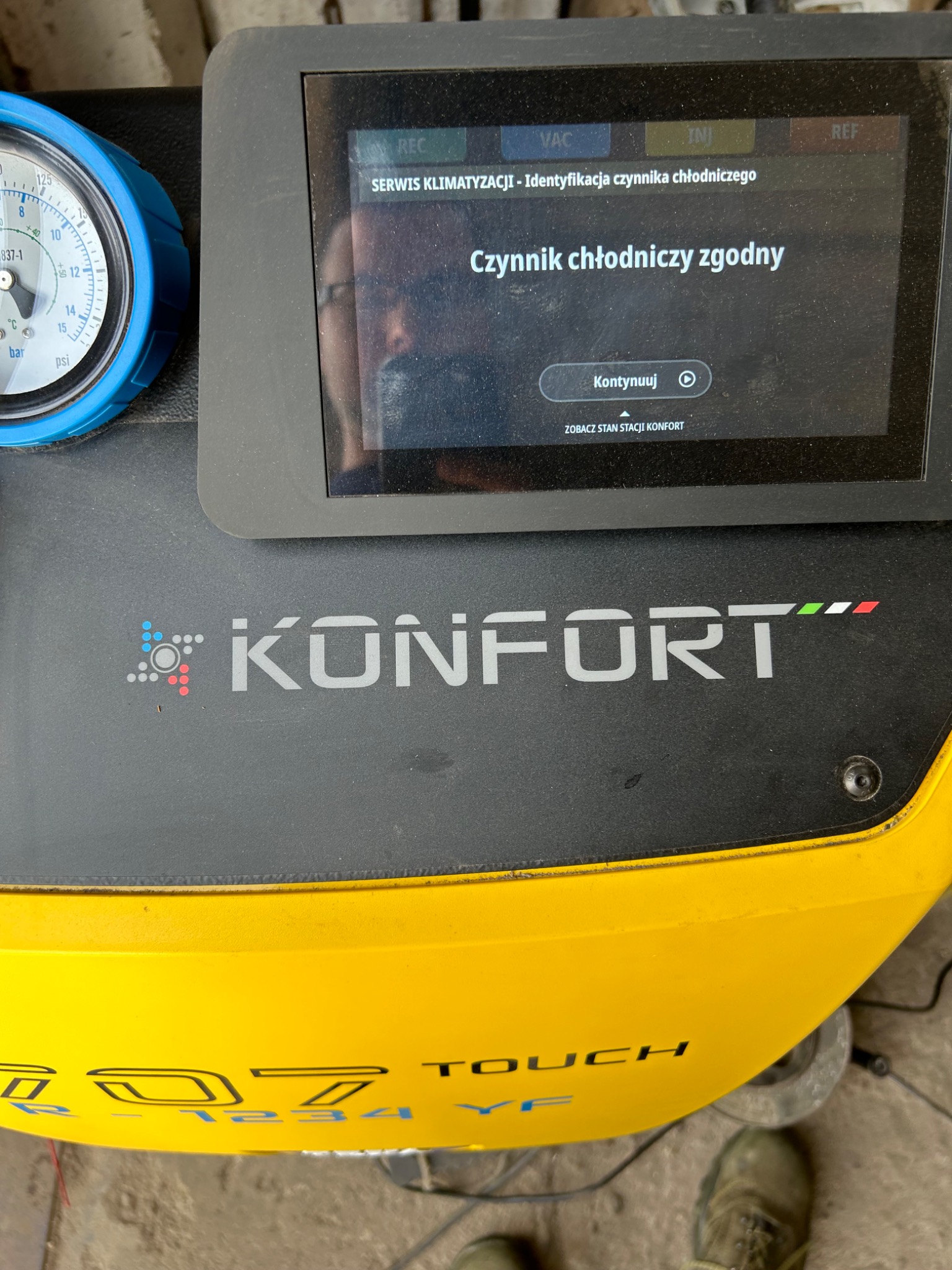 Urządzenie diagnostyczne Konfort z ekranem wyświetlającym komunikat 'Czynnik chłodniczy zgodny', widoczny manometr z niebieską obudową i żółta obudowa urządzenia z napisem 707 Touch.