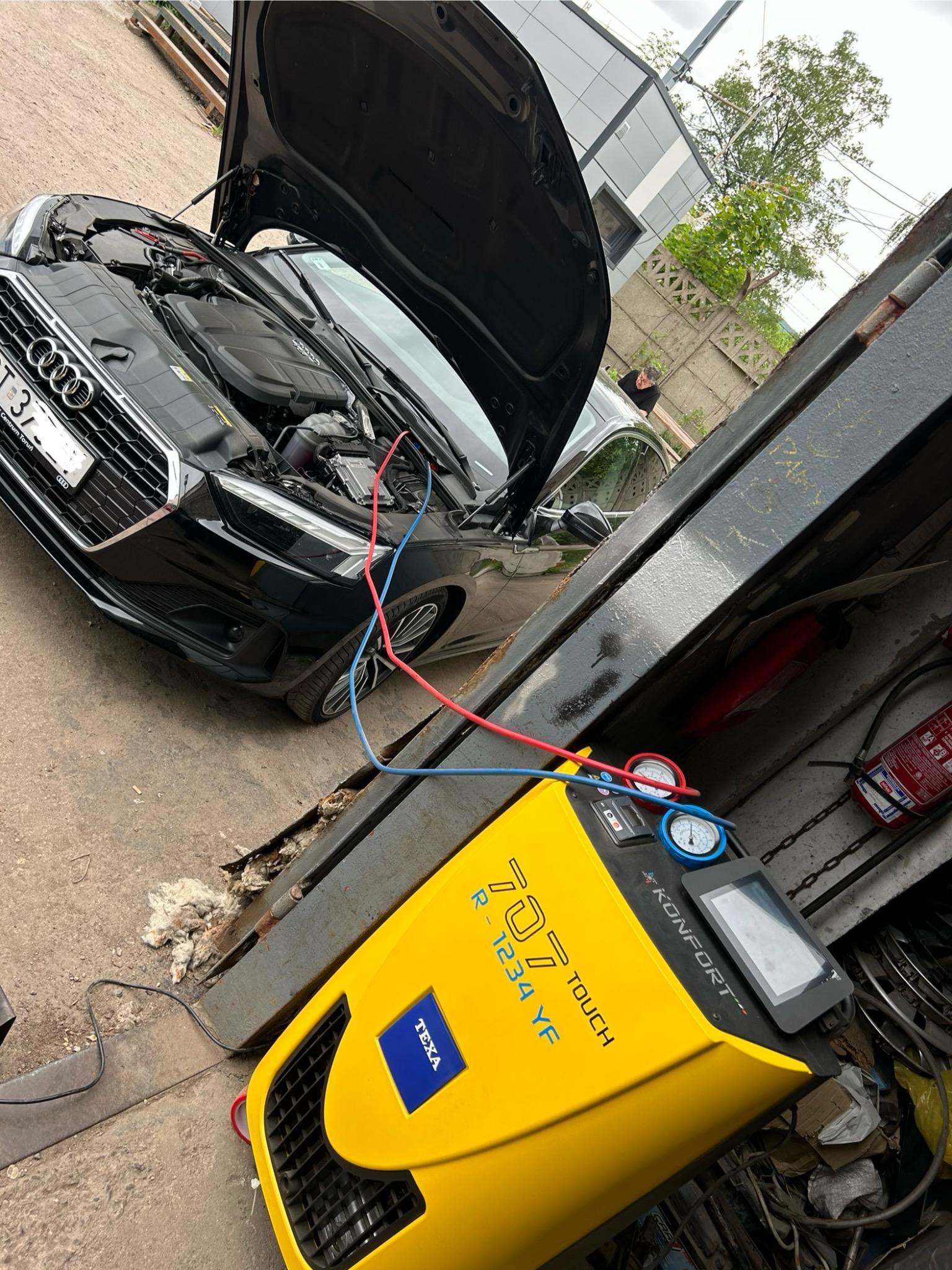 Czarny samochód marki Audi z otwartą maską, podłączony do żółtego urządzenia TEXA KONFORT 707 TOUCH R-1234 YF za pomocą czerwonych i niebieskich przewodów, podczas serwisu w warsztacie samochodowym.