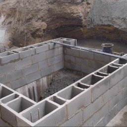 Łukasz Wleczyk - Fundament z bloczków betonowych w wykopie, poziomica i wiadro widoczne na górze, ziemia wokół wykopu.