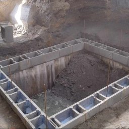 Łukasz Wleczyk - Fundament z bloczków betonowych w wykopie, widoczne pręty zbrojeniowe, ziemia i woda wewnątrz konstrukcji, ściany wykopu z jasnej ziemi.