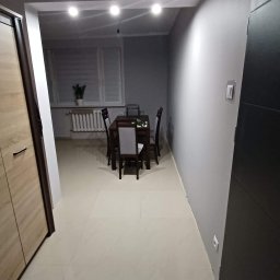 Łukasz Wleczyk - Jasne płytki podłogowe w szarym pokoju z ciemnymi meblami: stół z czterema krzesłami, grzejnik pod oknem z żaluzjami i szafa z jasnego drewna.