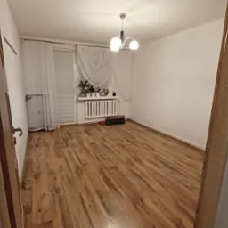 Pokój z ułożonymi panelami podłogowymi o wzorze drewna, widok zza uchylonych drzwi z ościeżnicą, na ścianie grzejnik pod oknem z firankami.