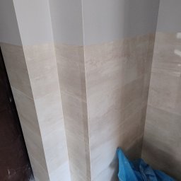 Łukasz Wleczyk - Wykończona ściana łazienki z beżowych płytek imitujących kamień, widoczny fragment podłogi w trakcie prac i niebieski worek na odpady budowlane w rogu.