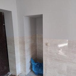 Łukasz Wleczyk - Świeżo pomalowane na szaro ściany łazienki z wykończonymi płytkami do połowy wysokości, z widocznym gniazdkiem elektrycznym i niebieskim workiem na śmieci budowlane w niszy.