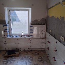 Łukasz Wleczyk - Świeżo ułożone, jasne płytki ścienne w trakcie fugowania, z widocznymi krzyżykami dystansowymi i niebieską taśmą malarską, okno z widokiem na zewnątrz, fragmenty nieotynkowanej ściany i żółte ślady...