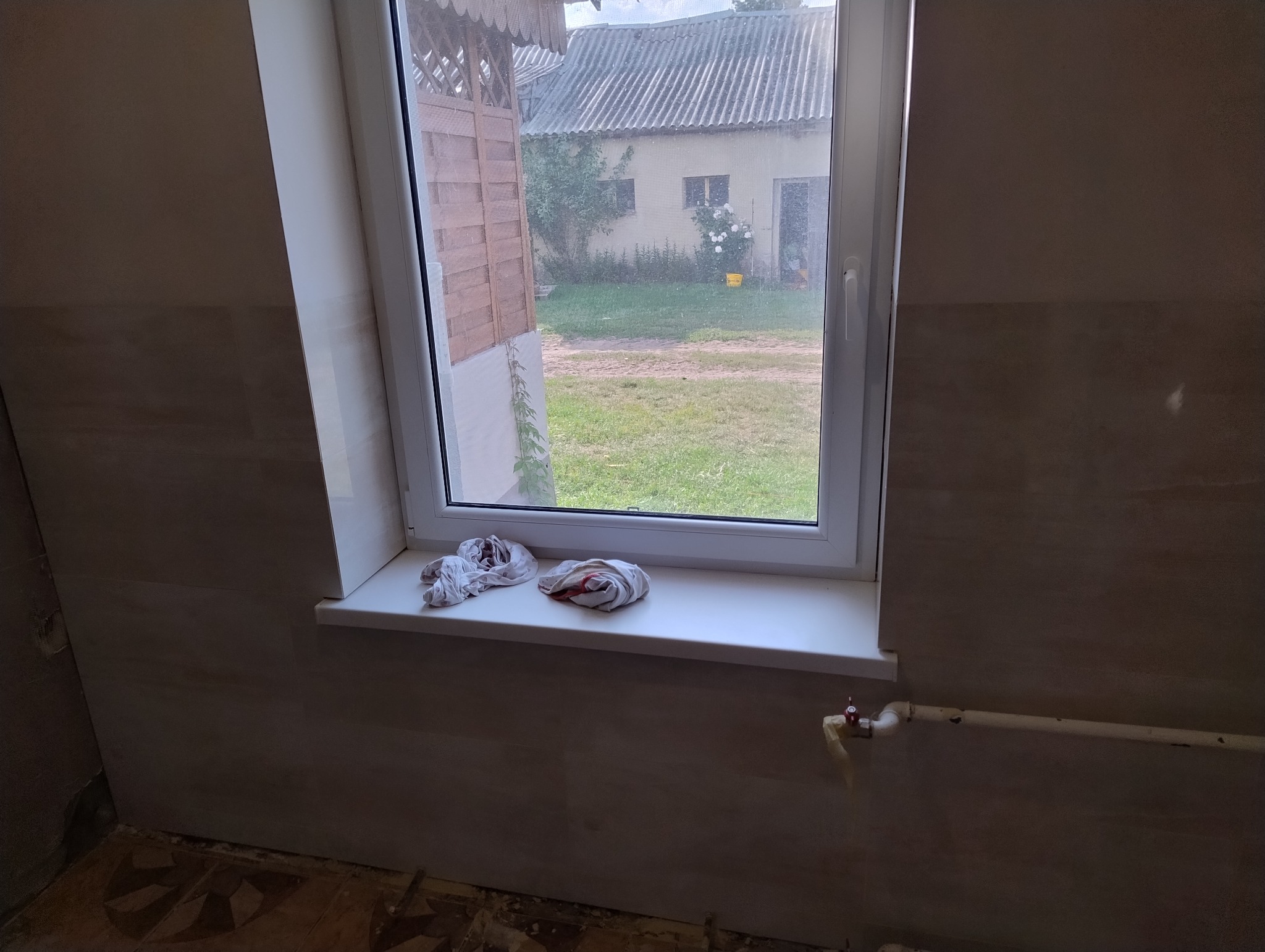 Ściana w trakcie remontu, wyłożona płytkami imitującymi kamień, z widocznym oknem, na parapecie leżą brudne ścierki, a poniżej wystaje rura z zaworem, podłoga częściowo odkryta.
