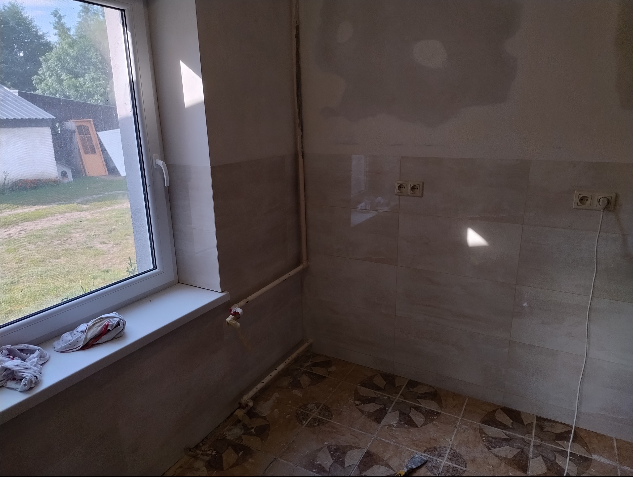 Remontowana łazienka z beżowymi płytkami ściennymi, częściowo zagruntowaną ścianą, starymi rurami, brudnymi szmatami na parapecie i podłogą z wzorem kwiatowym w trakcie wymiany płytek.