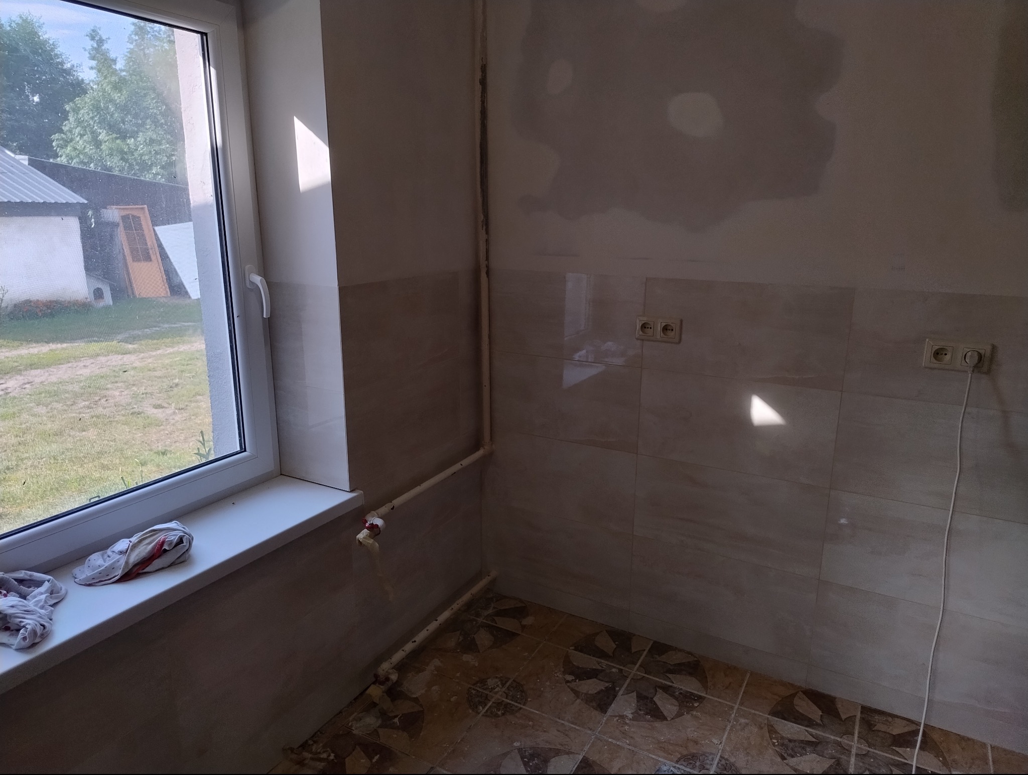 Fragment wnętrza w trakcie prac: ściany wyłożone płytkami, niedokończone gładzie na górze, widoczne rury, okno z widokiem na zewnątrz i szmatki na parapecie.