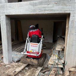 APL SERVICE SPÓŁKA Z OGRANICZONĄ ODPOWIEDZIALNOŚCIĄ - Kompaktowa minikoparka Takeuchi TB210R wewnątrz surowego budynku z betonu komórkowego, operator w czerwonej kurtce, widoczne deski i gruz na podłożu.