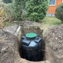 Czarny plastikowy zbiornik z zieloną pokrywą umieszczony w wykopie ziemnym, widoczne ślady świeżo wykonanych prac ziemnych, w tle zielona roślinność i fragment budynku.