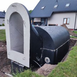 APL SERVICE SPÓŁKA Z OGRANICZONĄ ODPOWIEDZIALNOŚCIĄ - Prefabrykowany, betonowy zbiornik szamba z kominem inspekcyjnym, częściowo zagłębiony w wykopie na trawiastym terenie, na tle domu z ciemną dachówką