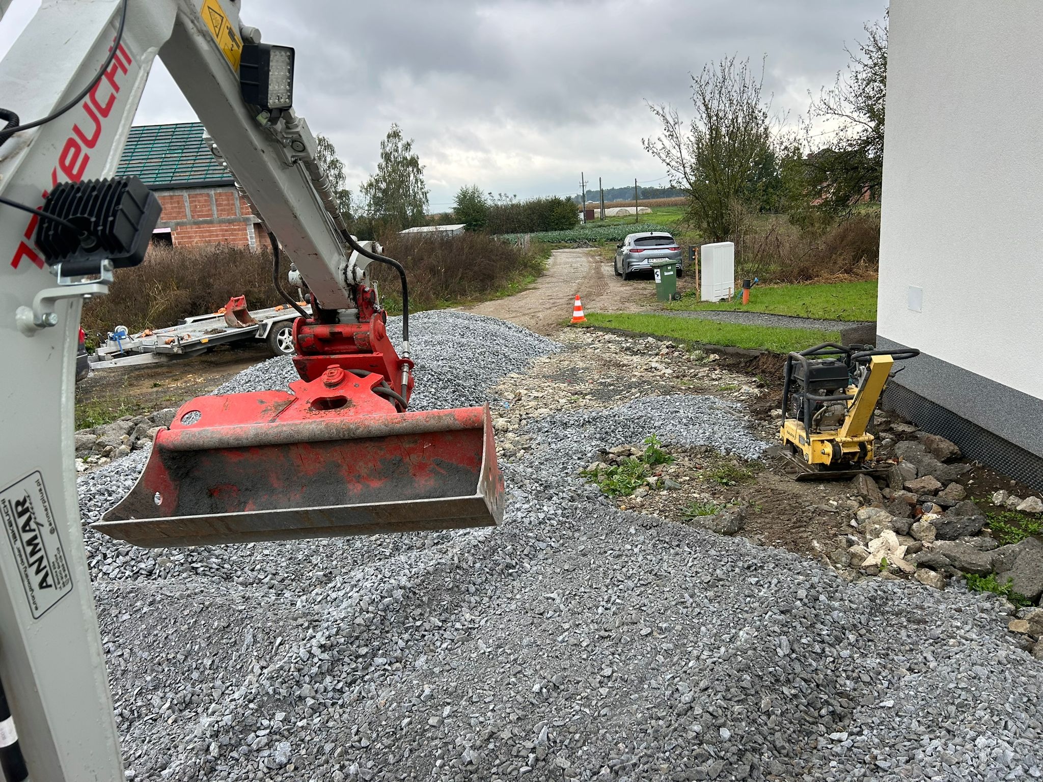 Mini koparka z brudną łyżką nad stertą szarego gruzu, obok zagęszczarka wibracyjna, w tle dom w budowie i polna droga