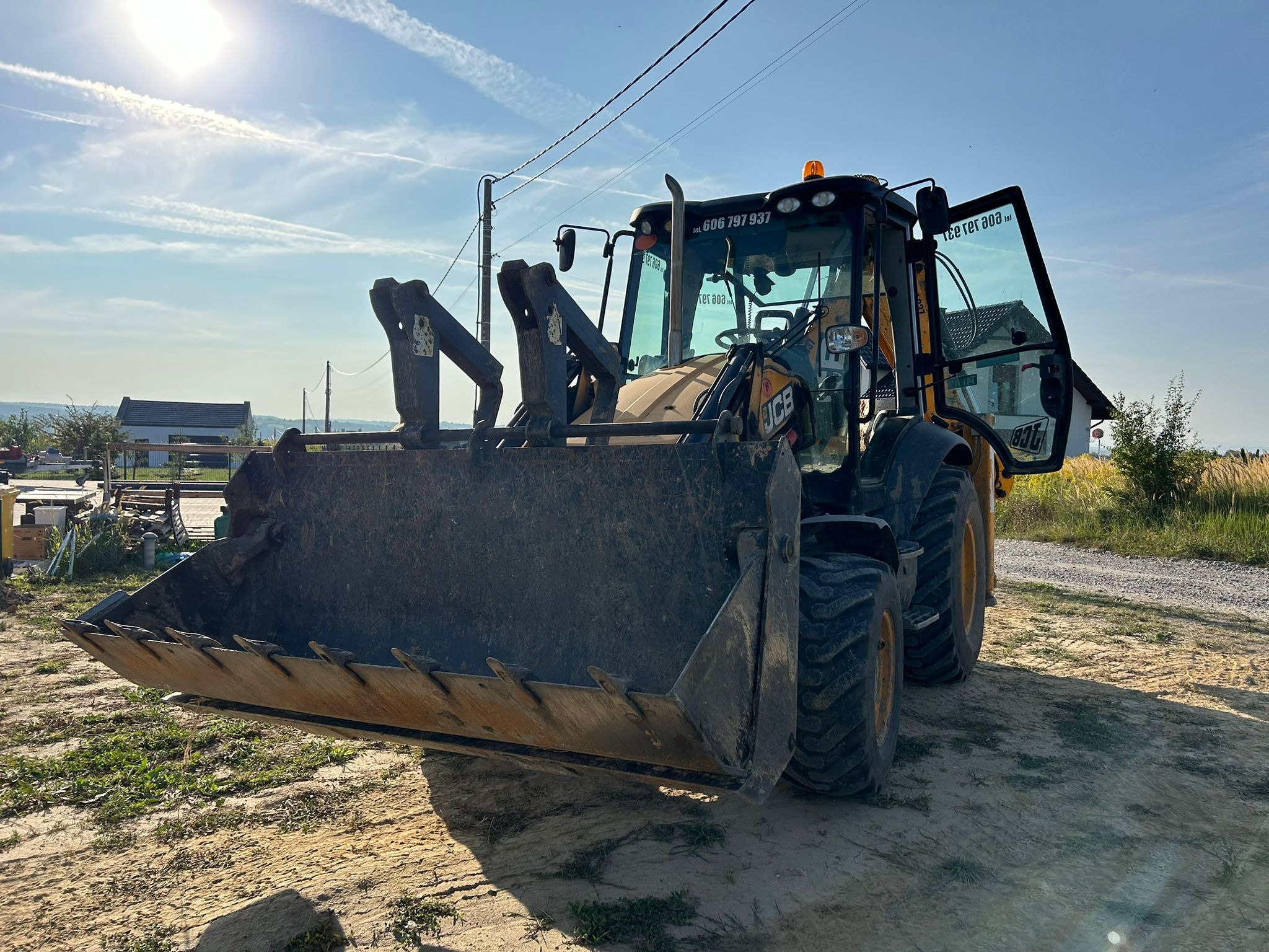 Żółto-czarna koparko-ładowarka JCB stoi na piaszczystym terenie z otwartymi drzwiami kabiny, widoczne budynki w tle pod błękitnym niebem z chmurami.
