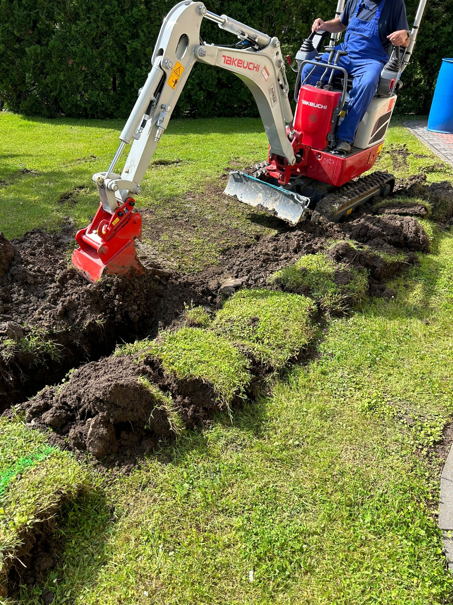 Mini koparka Takeuchi z czerwoną łyżką w trakcie wykonywania wąskiego wykopu w trawniku, widoczne odcięte fragmenty darni.