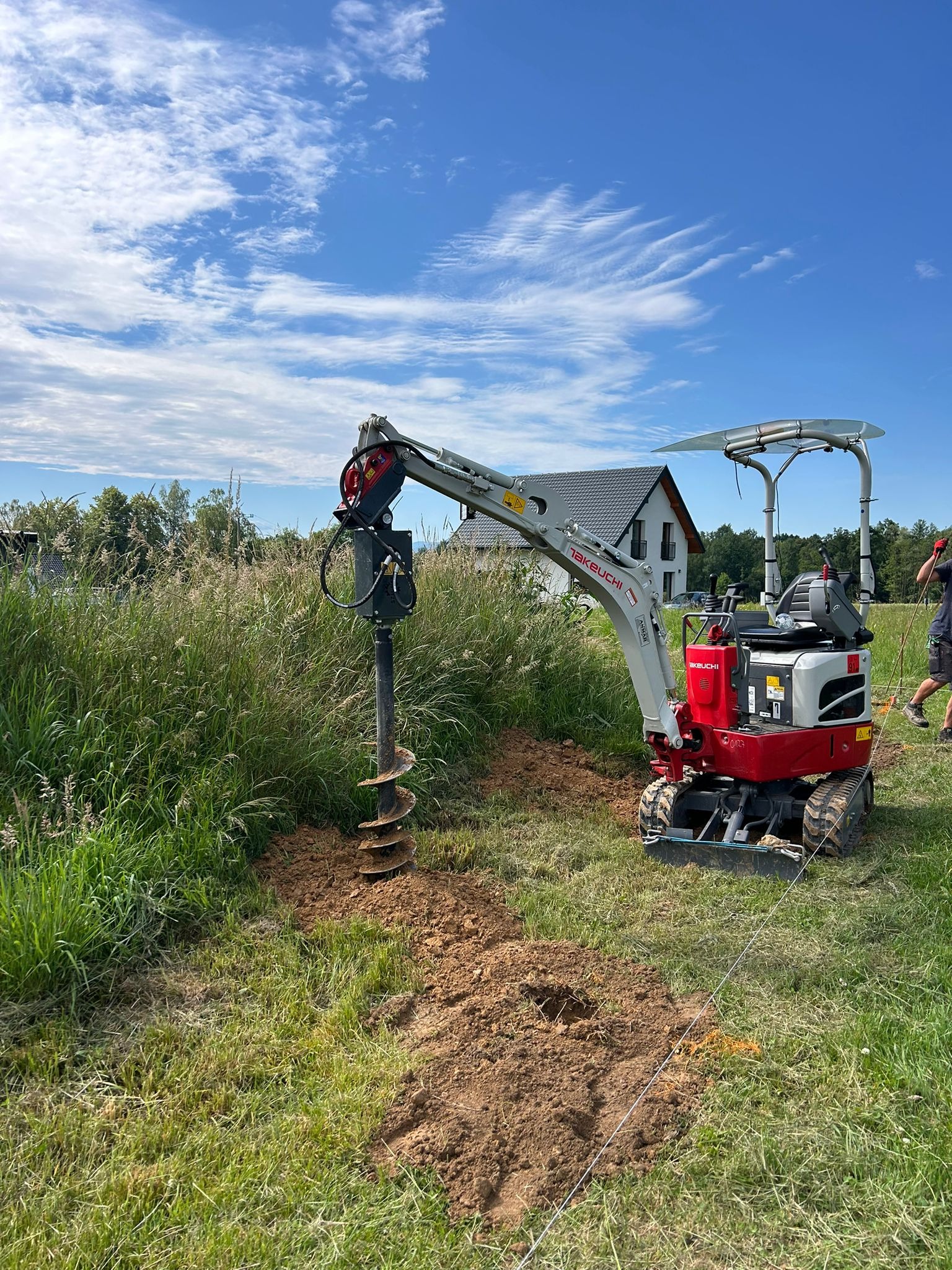 Czerwono-biała minikoparka Takeuchi z wiertnicą świdrową w trakcie wiercenia otworu w ziemi na trawiastym terenie, widoczna linia wyznaczająca trasę wiercenia, w tle dom i błękitne niebo z chmurami.