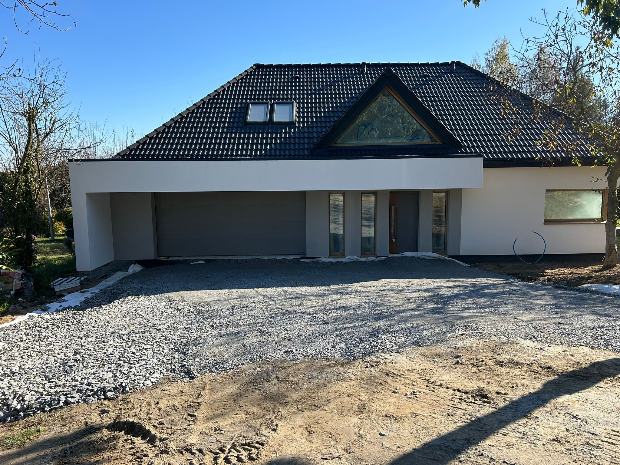 Nowoczesny dom z czarnym dachem i szarą bramą garażową, podjazd wysypany szarym żwirem, widoczne ślady opon na ziemi.