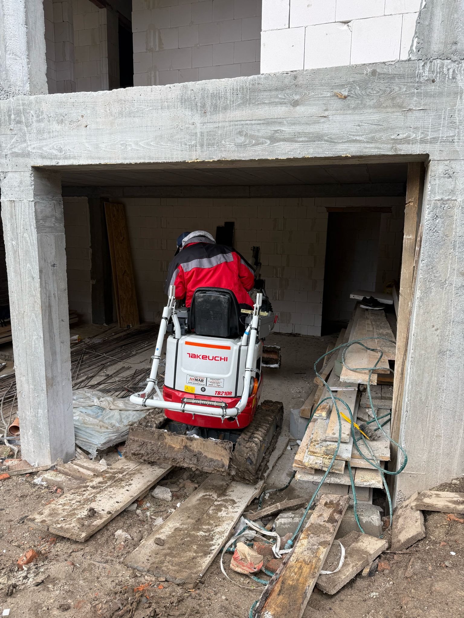Kompaktowa minikoparka Takeuchi TB210R wewnątrz surowego budynku z betonu komórkowego, operator w czerwonej kurtce, widoczne deski i gruz na podłożu.