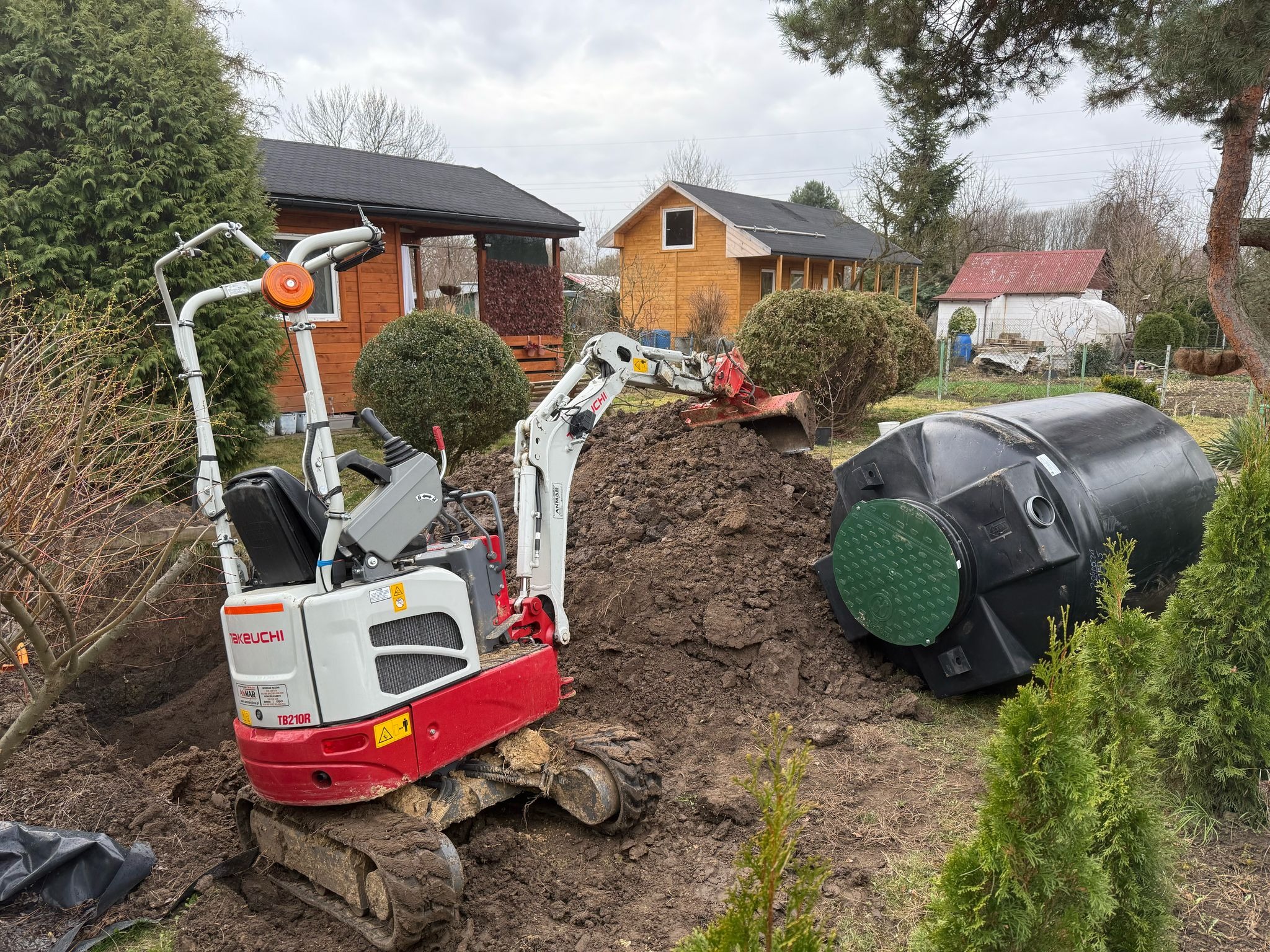 Mini koparka Takeuchi TB210R wykopuje ziemię obok czarnego zbiornika z zieloną pokrywą na tle domów i zieleni.