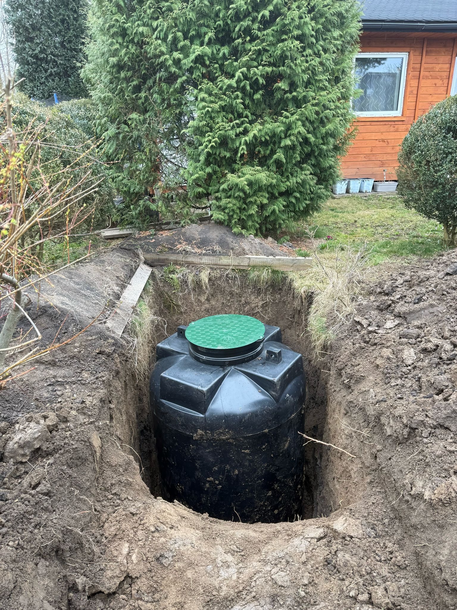 Czarny plastikowy zbiornik z zieloną pokrywą umieszczony w wykopie ziemnym, widoczne ślady świeżo wykonanych prac ziemnych, w tle zielona roślinność i fragment budynku.