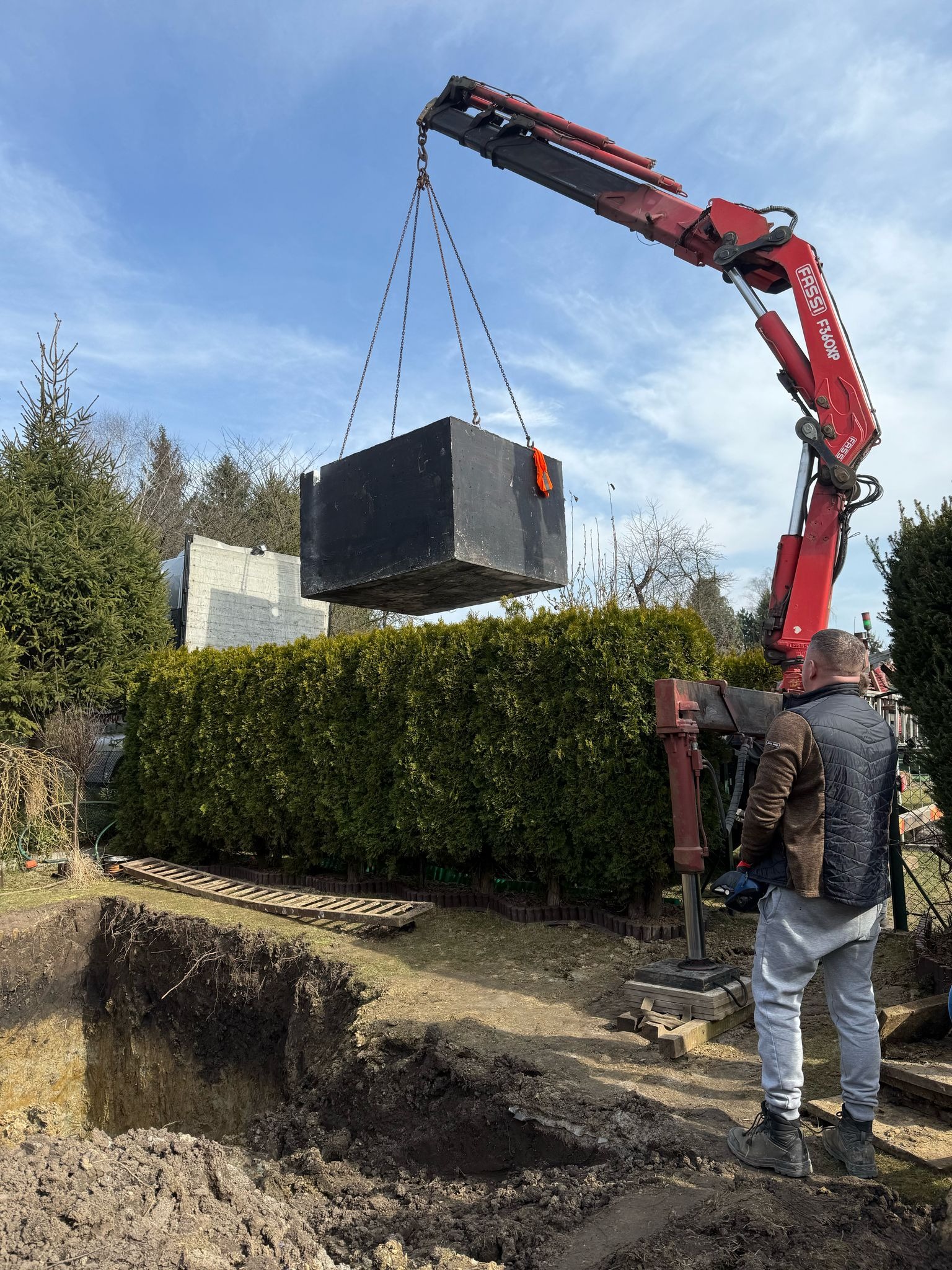 Ciężki, betonowy element szamba zawieszony na łańcuchach dźwigu Fassi F340XP, opuszczany do wykopu w ziemi, nadzorowany przez mężczyznę w szarych spodniach i czarnej kamizelce na tle zielonego...