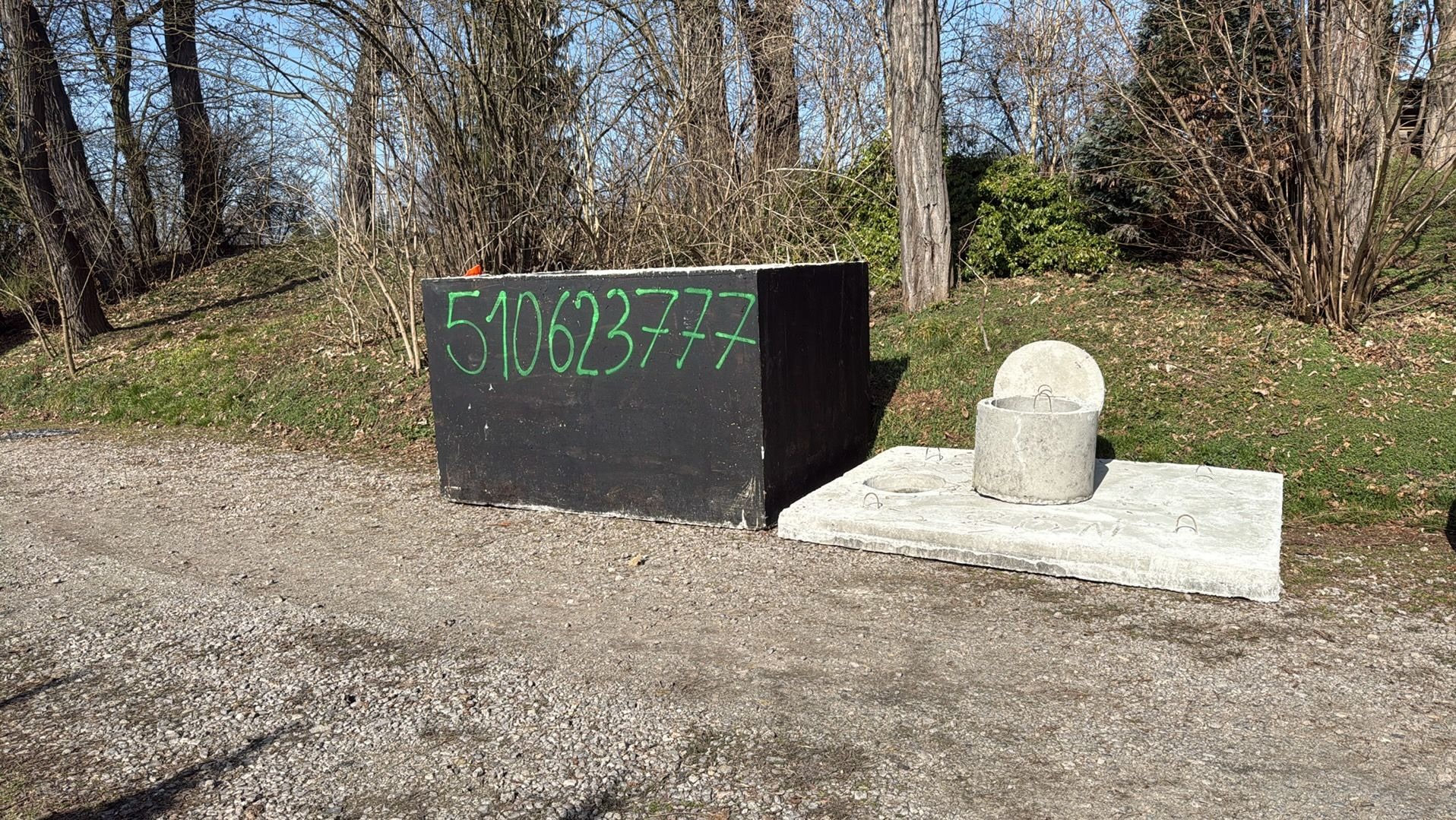 Betonowy zbiornik szamba z napisanym zielonym markerem numerem telefonu, umieszczony obok betonowej płyty z włazem, na żwirowej powierzchni w pobliżu drzew.