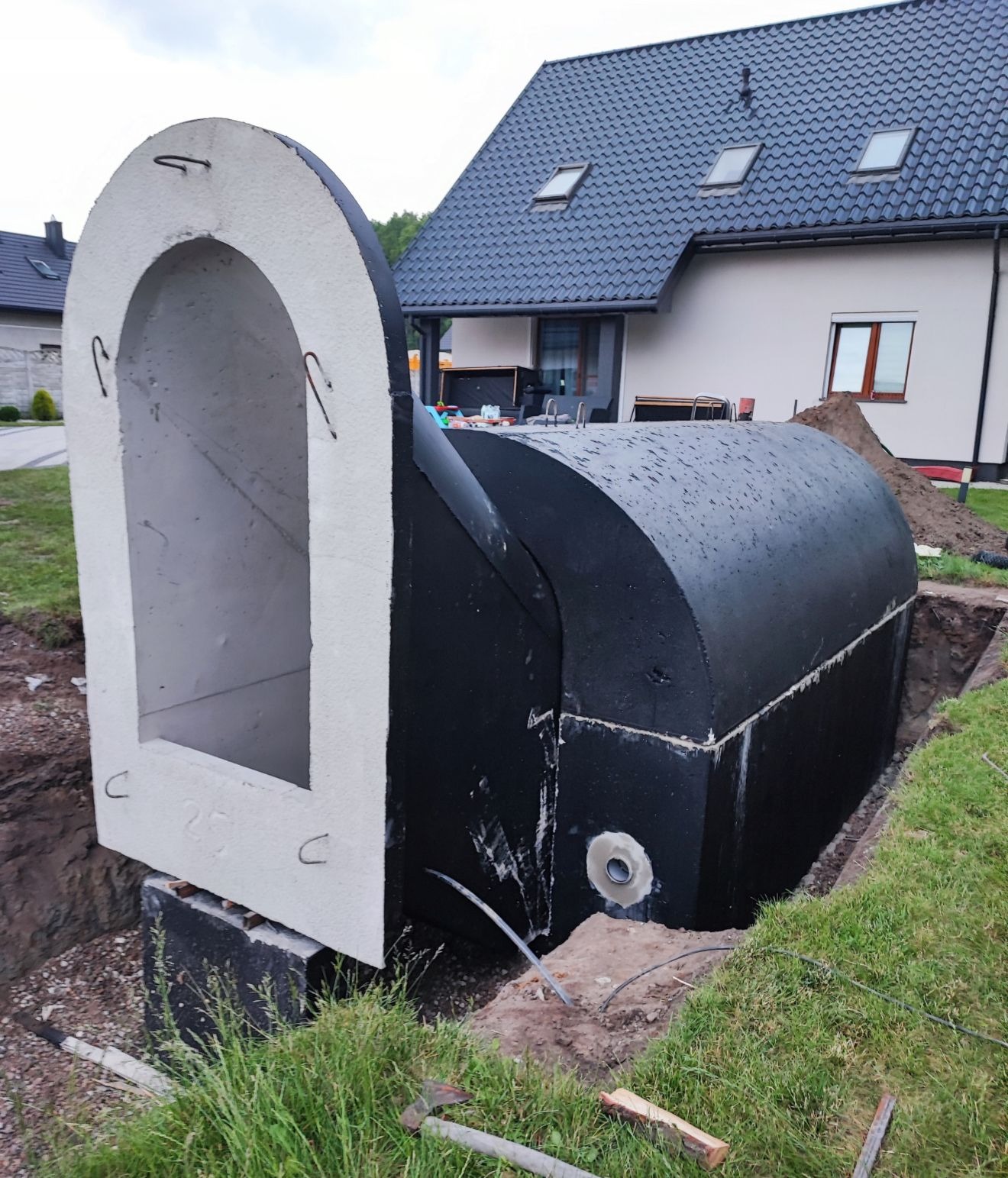 Prefabrykowany, betonowy zbiornik szamba z kominem inspekcyjnym, częściowo zagłębiony w wykopie na trawiastym terenie, na tle domu z ciemną dachówką
