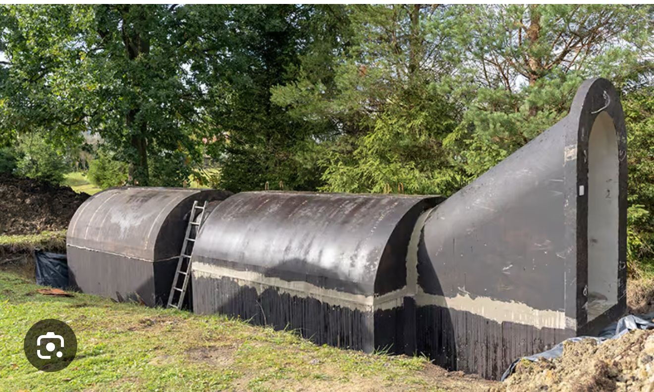 Montaż modułowego zbiornika podziemnego z włókna szklanego, widoczna drabina oparta o zbiornik, ziemia wokół wykopu, drzewa w tle.