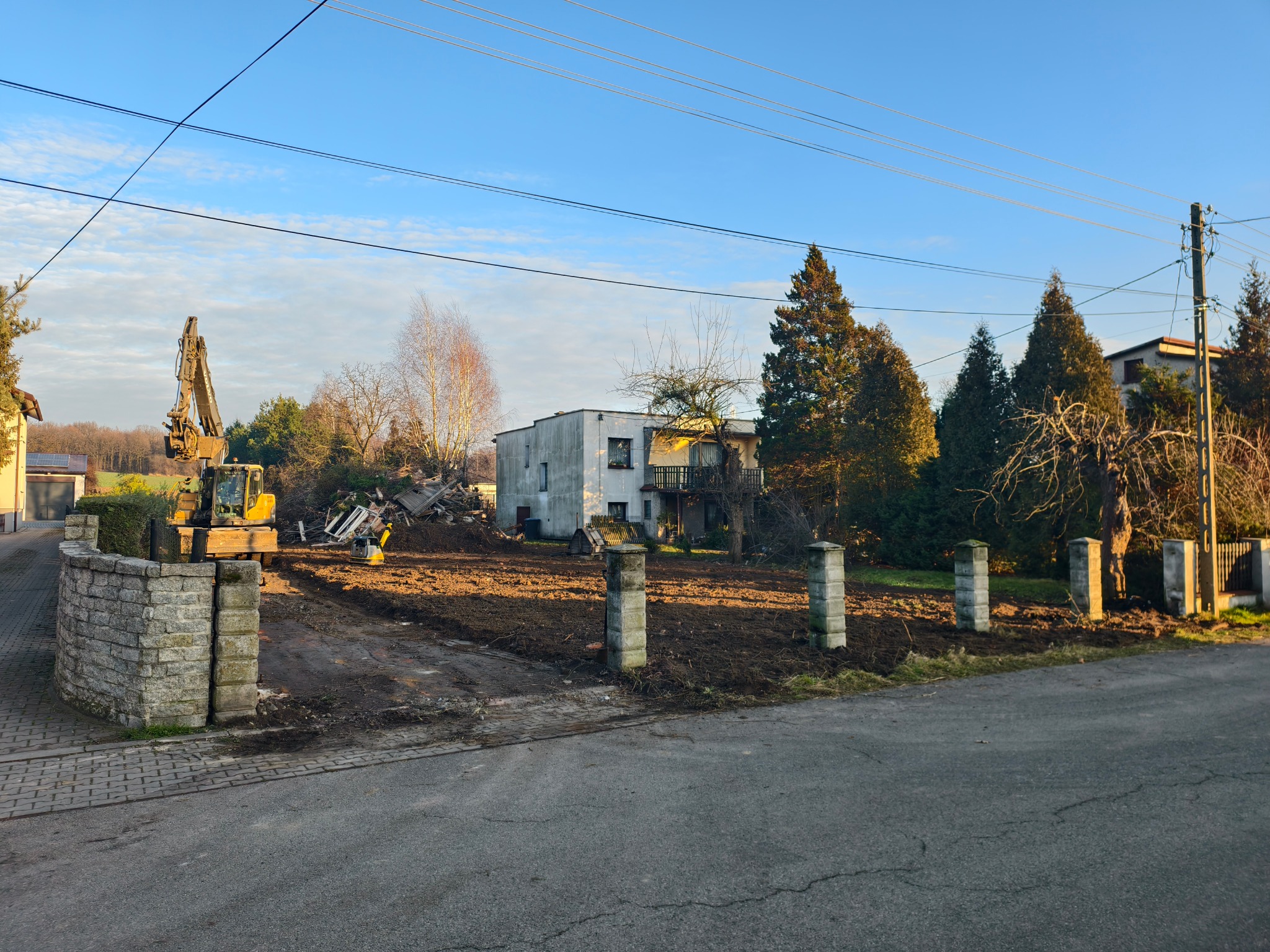 Żółta koparka na działce po rozbiórce domu, z gruzem i fundamentami w tle. Widoczne słupki ogrodzeniowe i droga dojazdowa. Błękitne niebo.