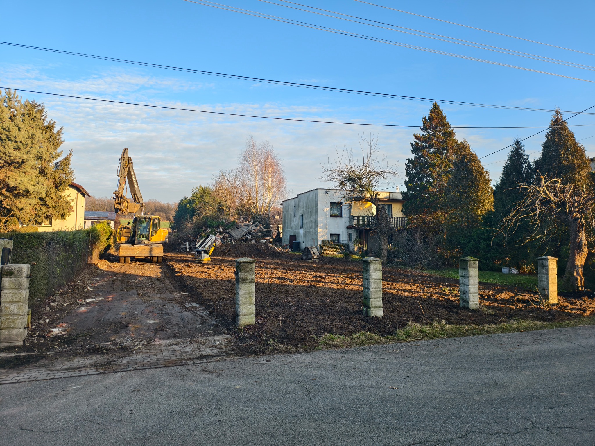 Żółta koparka na działce po wyburzeniu, przygotowuje teren pod budowę. Widoczne gruzy i resztki roślinności. W tle budynek mieszkalny i błękitne niebo.