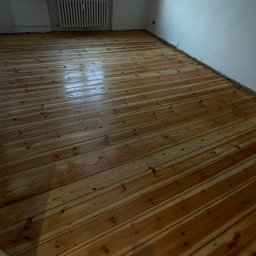 Mateusz Wardaszewski - Świeżo wycyklinowana, lakierowana podłoga z desek w pustym pokoju z grzejnikiem pod oknem.